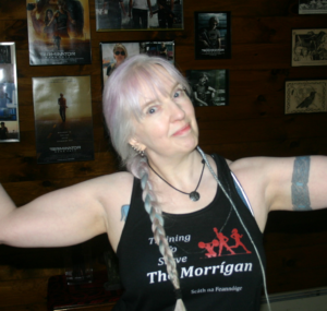 me goofing off in my gym: I’m a gray (some purple and blue tones) haired middle-aged woman with tattoos wearing a “Training to Serve The Morrígan” tank top